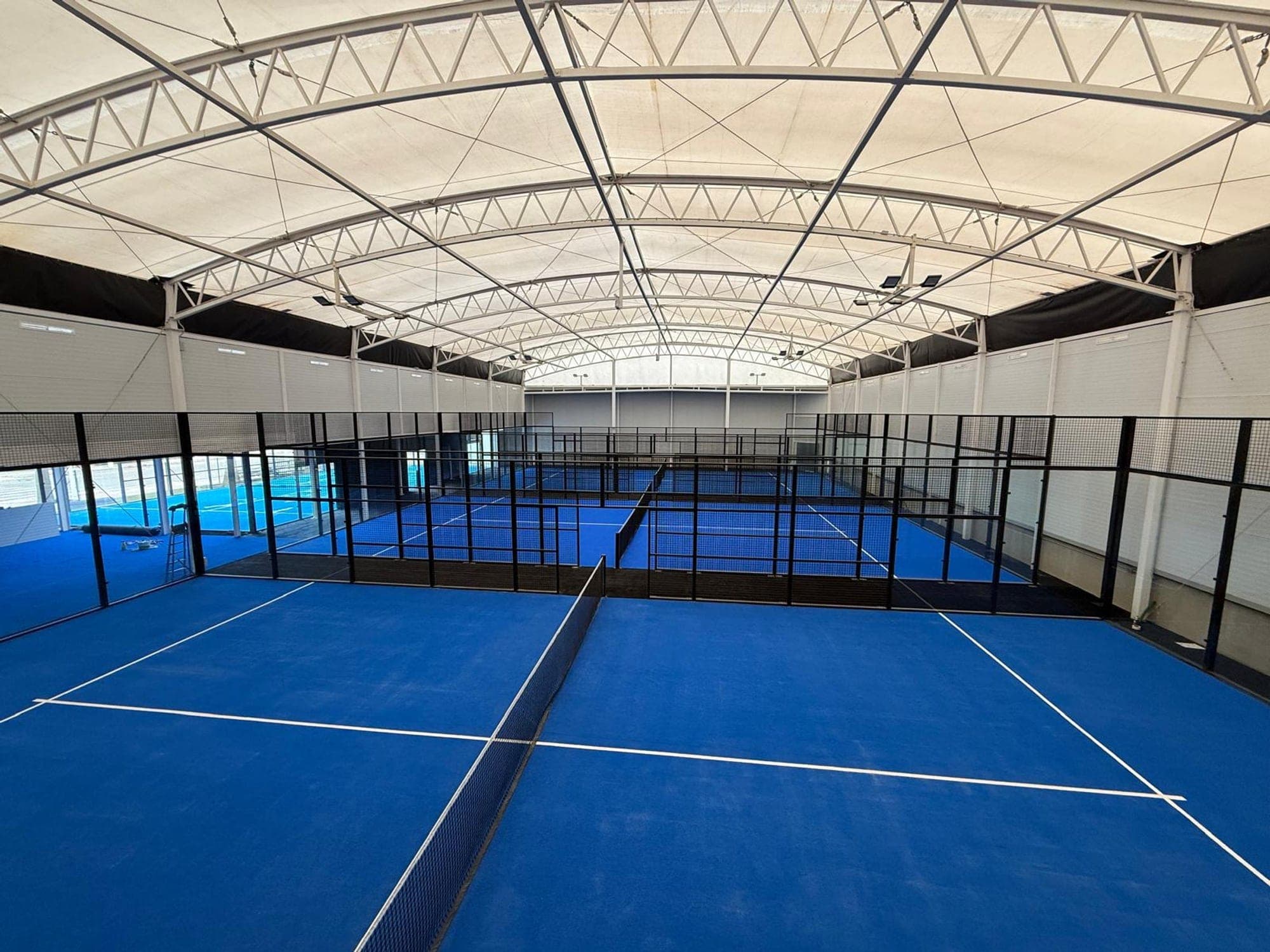 Interior de la nave de pádel del club, con las pistas indoor de césped azul bajo la lona y las cerchas blancas